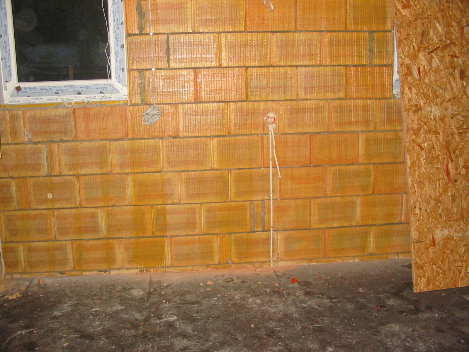 Ściana z pustaków ceramicznych z widocznymi przewodami elektrycznymi wychodzącymi ze ściany, okno w tle. Etap prac instalacyjnych w surowym stanie.