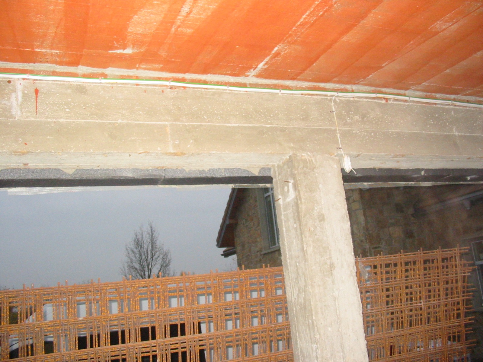 Surowy betonowy strop z wystającą kolumną i siatką zbrojeniową. Widok na budynek w tle przez otwór w konstrukcji. W tle widoczne przewody elektryczne.