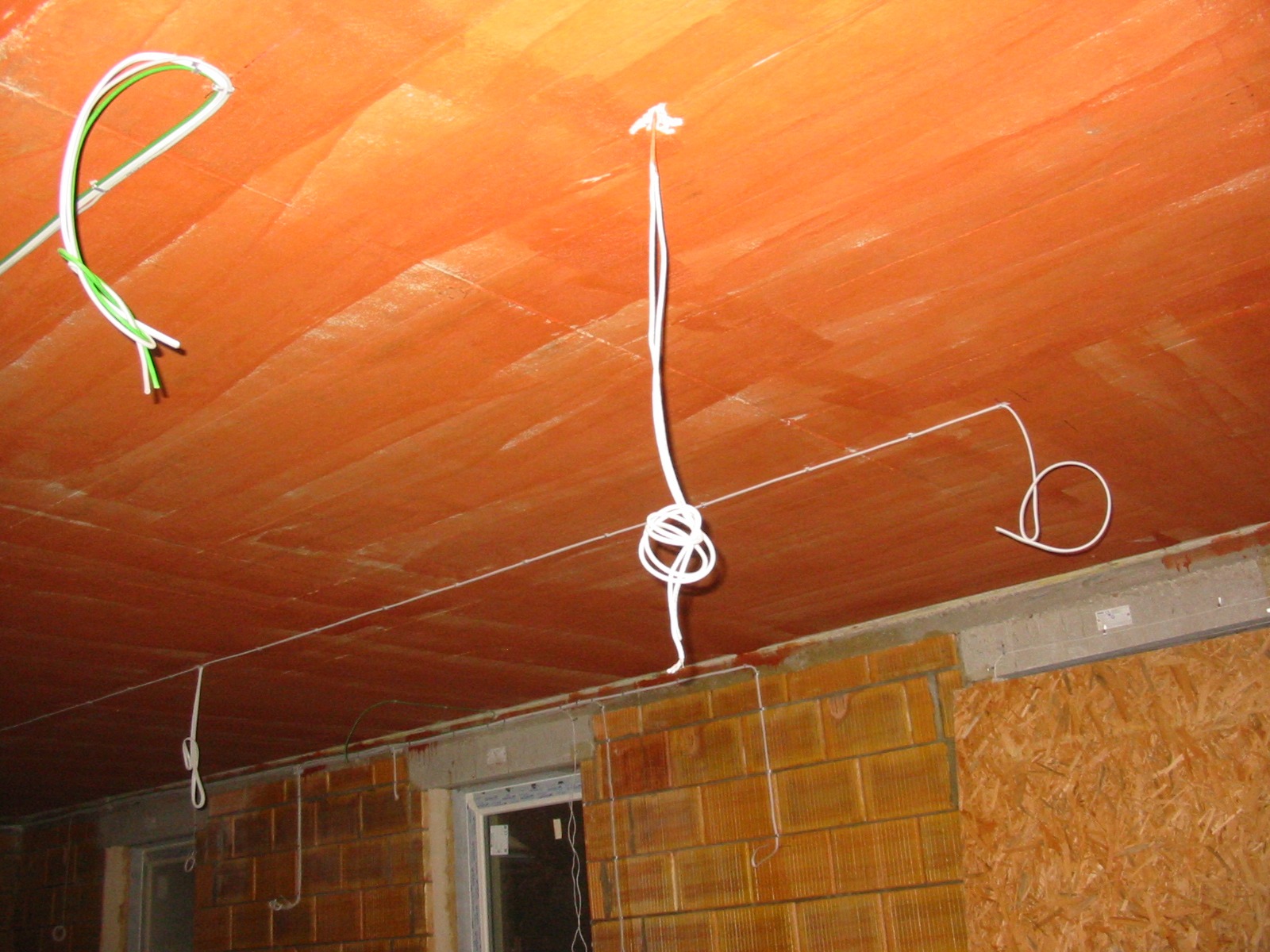 Instalacja elektryczna w surowym wnętrzu: przewody zwisają z pomarańczowego sufitu, widoczne okno i ściany z cegły oraz płyty OSB. Etap prac przed wykończeniem.