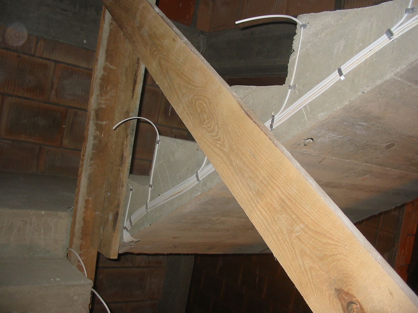 Surowy strop z widocznymi przewodami elektrycznymi, przymocowanymi klamrami. Drewniane stemple podtrzymują konstrukcję. Ceglany mur w tle.