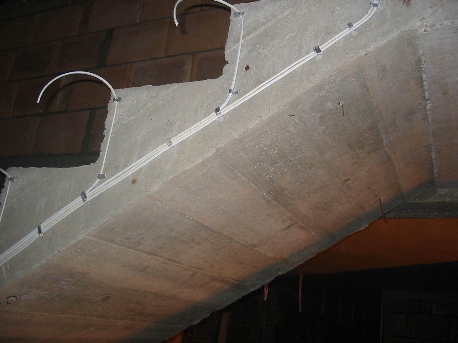 Surowy betonowy strop z przymocowanymi białymi rurkami instalacyjnymi za pomocą metalowych uchwytów. Widoczne fragmenty muru z cegły. Ujęcie z dołu.