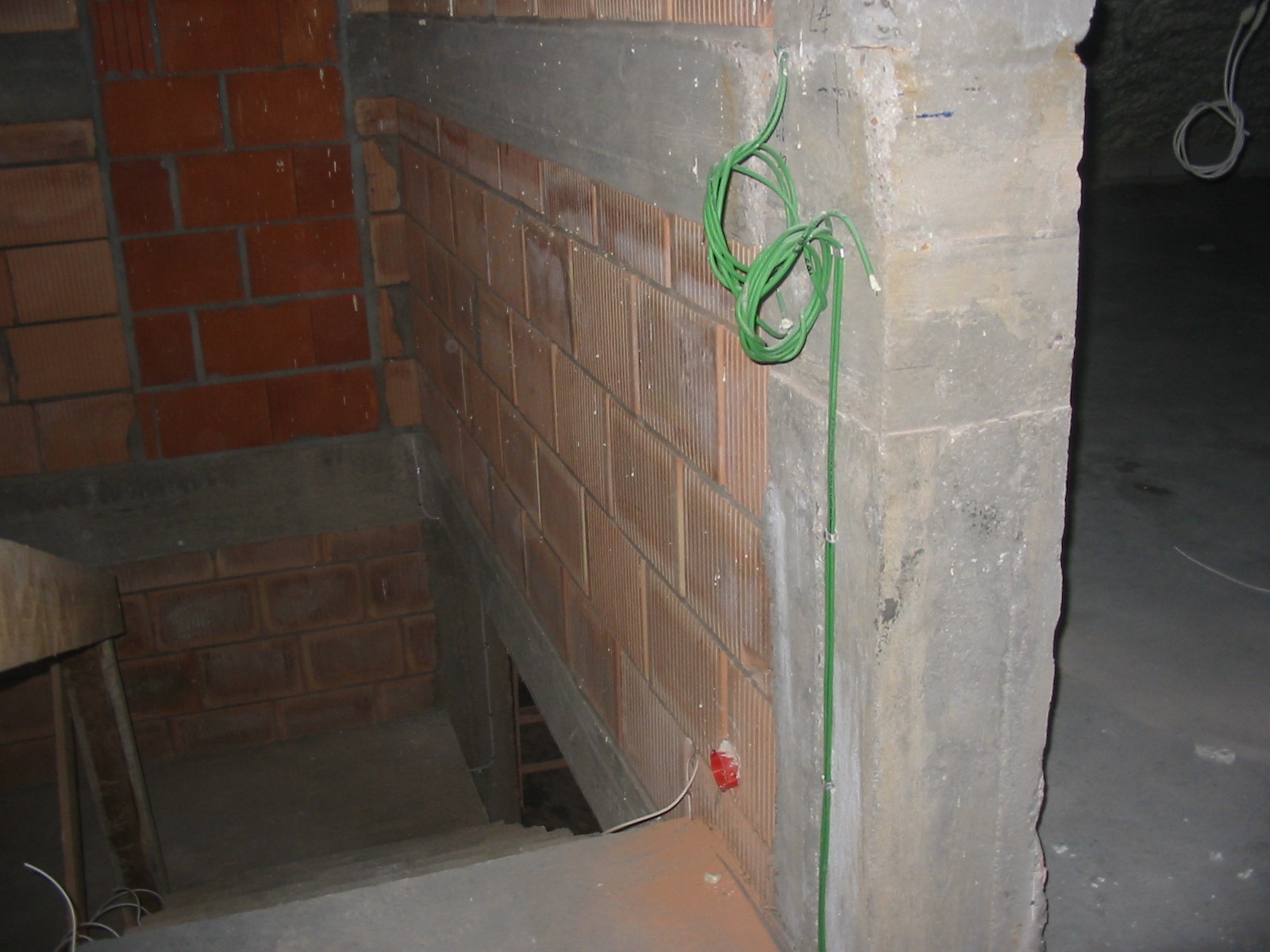 Surowa ściana z cegły z zielonymi kablami elektrycznymi przymocowanymi do ściany. Widoczna puszka elektryczna. W tle schody w stanie surowym.