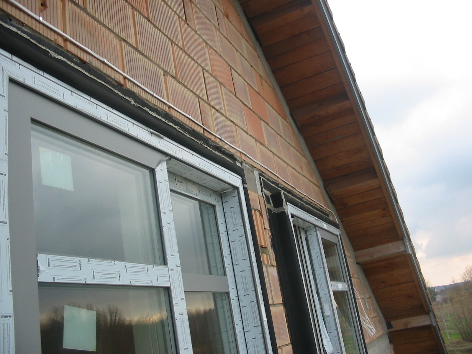 Montaż okien w budynku z cegły. Widoczne szare ramy okienne z folią ochronną, fragment dachu z drewnianą konstrukcją. Ujęcie z dołu, podkreślające wysokość budynku.