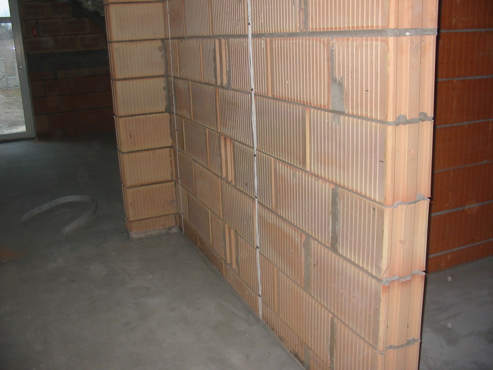 Ściana z pustaków ceramicznych Porotherm, widoczne spoiny i pionowe rowki. Szary betonowy podkład. W tle widoczne kolejne pustaki i fragment okna.