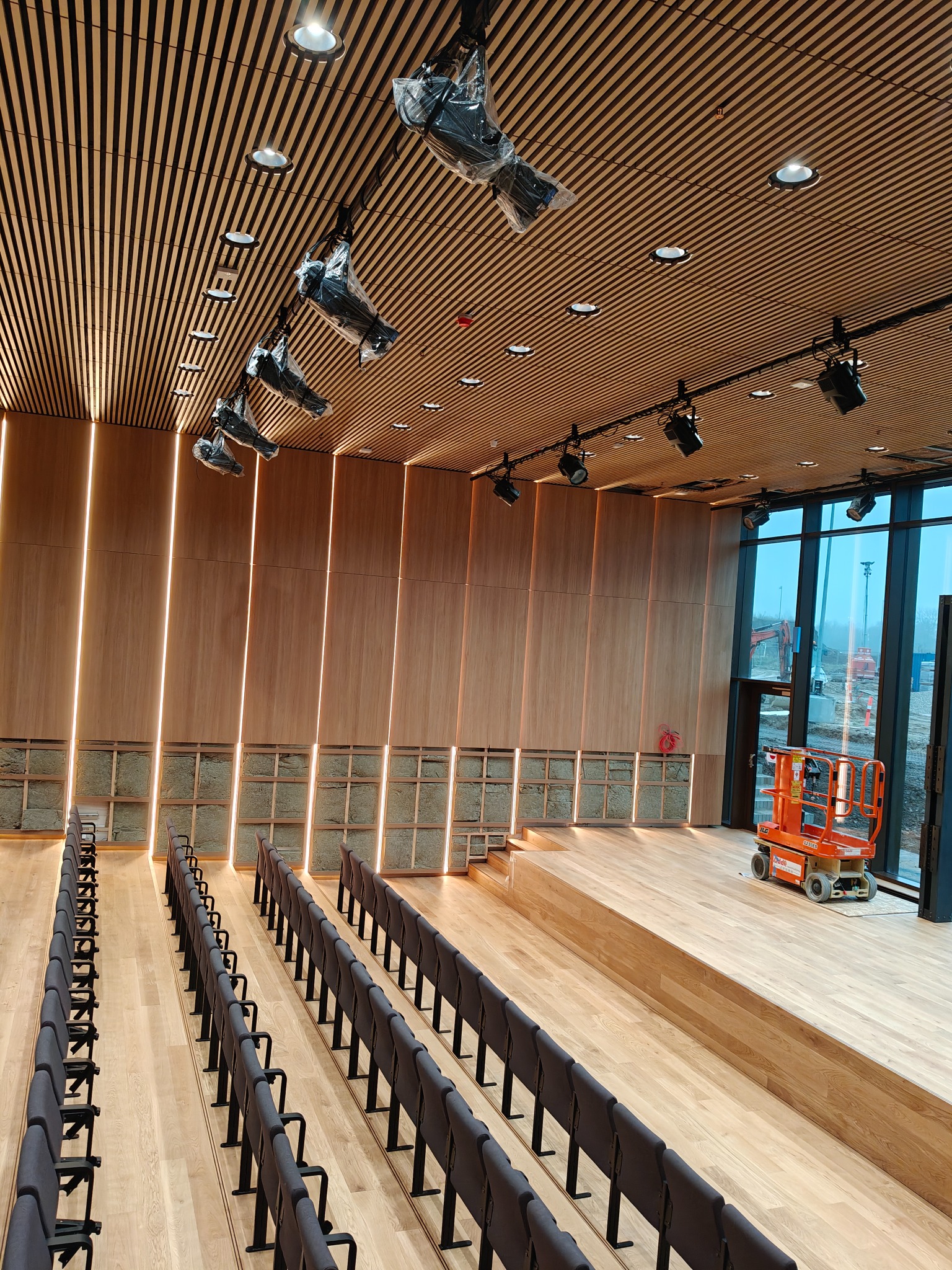 Sala widowiskowa w trakcie montażu oświetlenia: reflektory pod sufitem, podświetlane panele ścienne, rzędy krzeseł, podnośnik koszowy przy oknie.