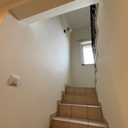 Jarek Lachowski - Klatka schodowa z beżowymi płytkami, widok z dołu. Na pierwszym planie para brązowych butów. Ściany w jasnym odcieniu beżu, okno na końcu schodów.