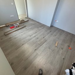 Jarek Lachowski - Układanie paneli podłogowych w jasnym pokoju. Widoczne narzędzia, paczki paneli i fragment osoby w roboczym ubraniu. Częściowo ułożona podłoga.