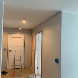 Jarek Lachowski - Przygotowanie do malowania: zabezpieczone taśmą ściany, drzwi i podłoga. Widoczna drabina, wałek malarski i folia ochronna. Ściany częściowo pomalowane na szaro.