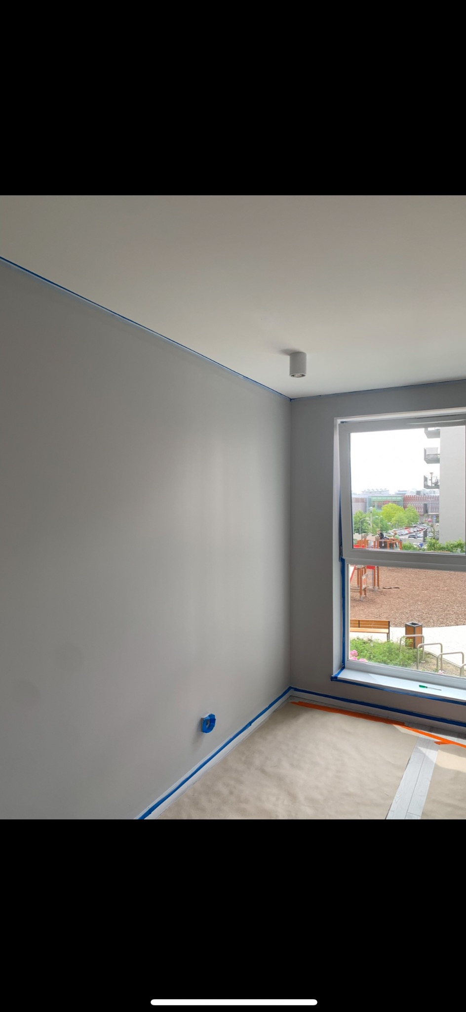 Pokój przygotowany do malowania: szare ściany, zabezpieczone taśmą malarską krawędzie, okno z widokiem na plac zabaw. Widoczna osłona gniazdka elektrycznego.