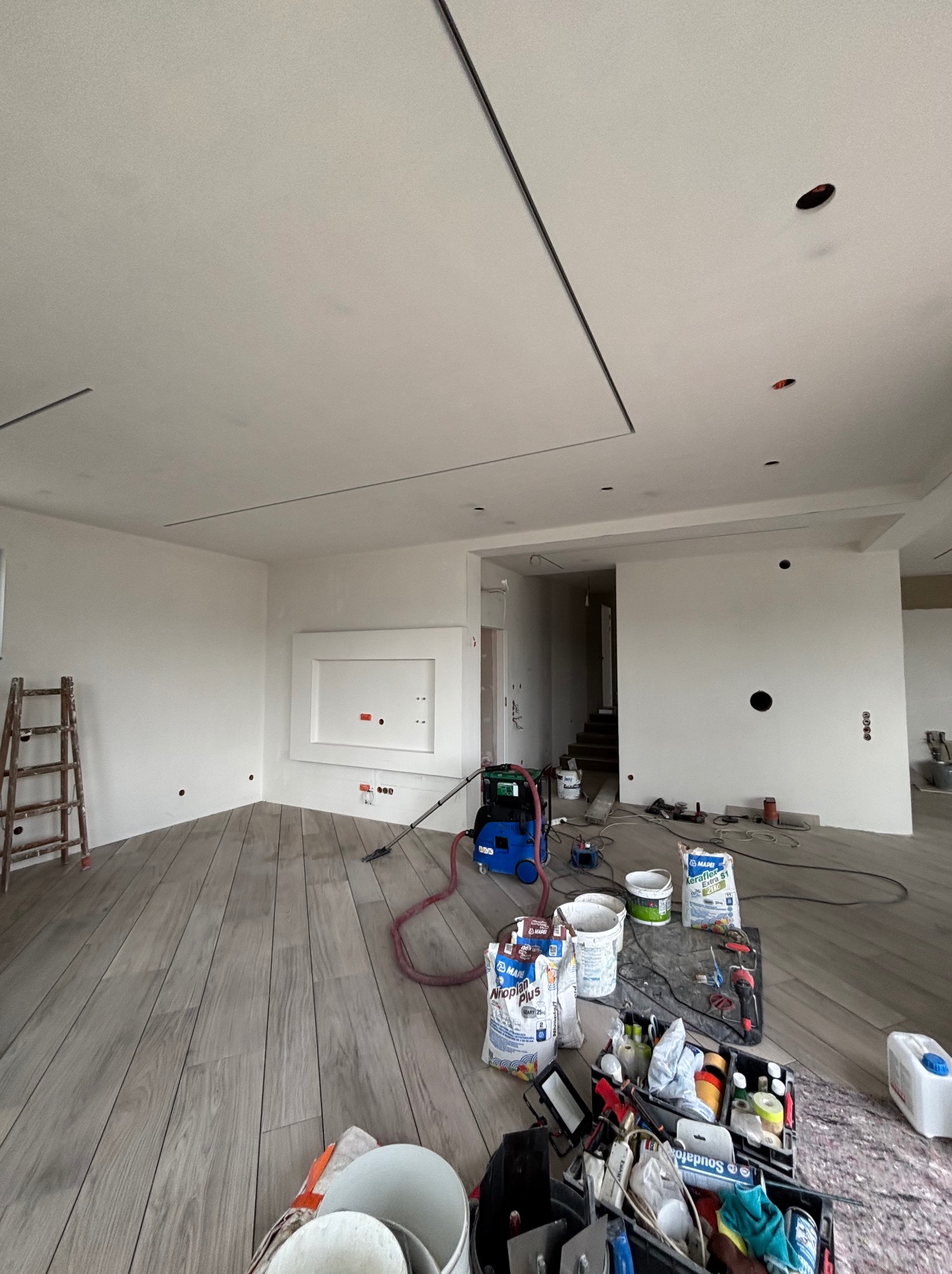 Wnętrze w trakcie prac wykończeniowych: gładkie, białe ściany, drewnopodobna podłoga, widoczne narzędzia i materiały budowlane, sufit z minimalistycznym oświetleniem.