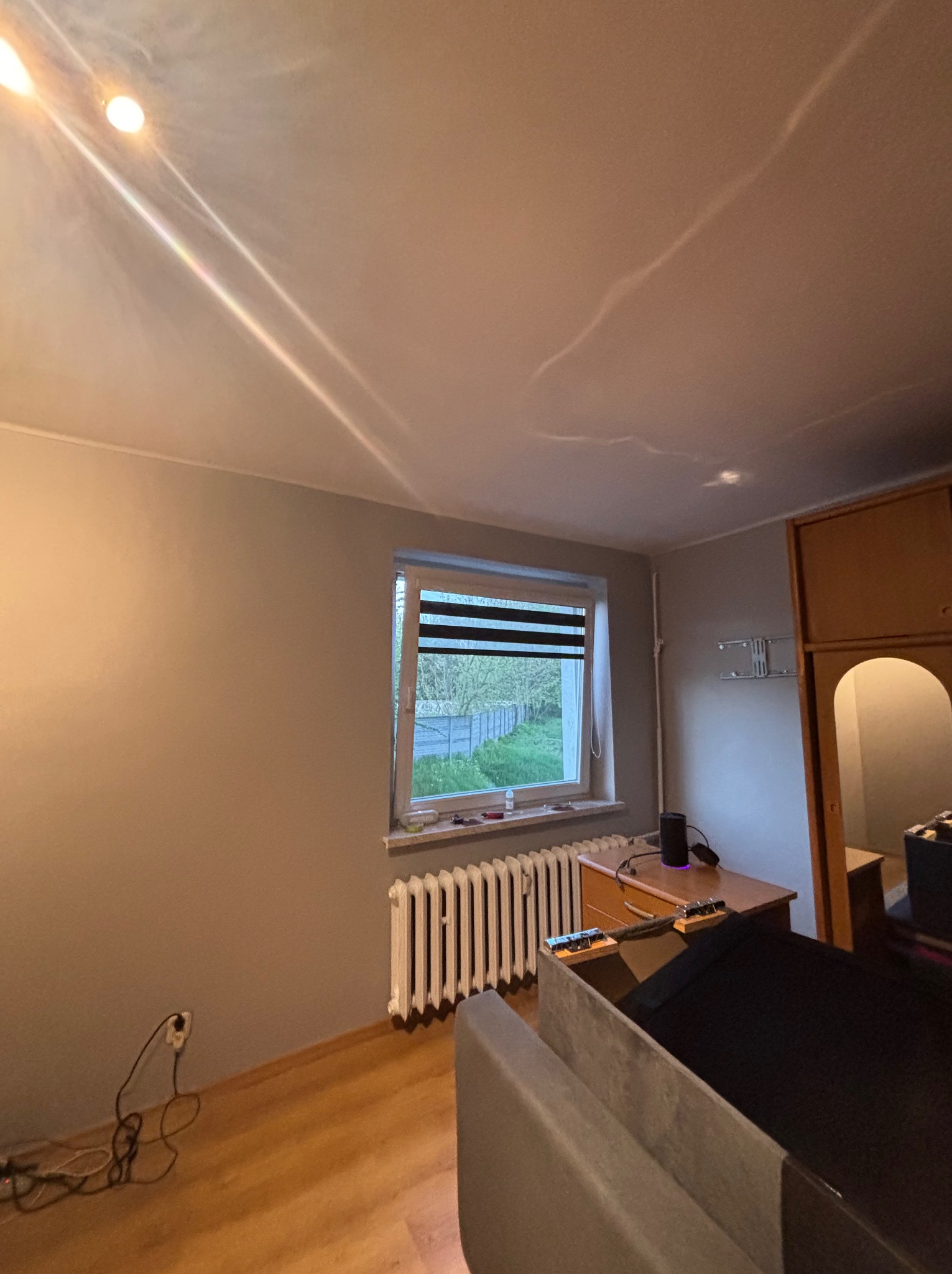 Pokój po malowaniu ścian na szaro, z oknem z żaluzjami, grzejnikiem, biurkiem i szafą. Widoczne ślady po remoncie na suficie.