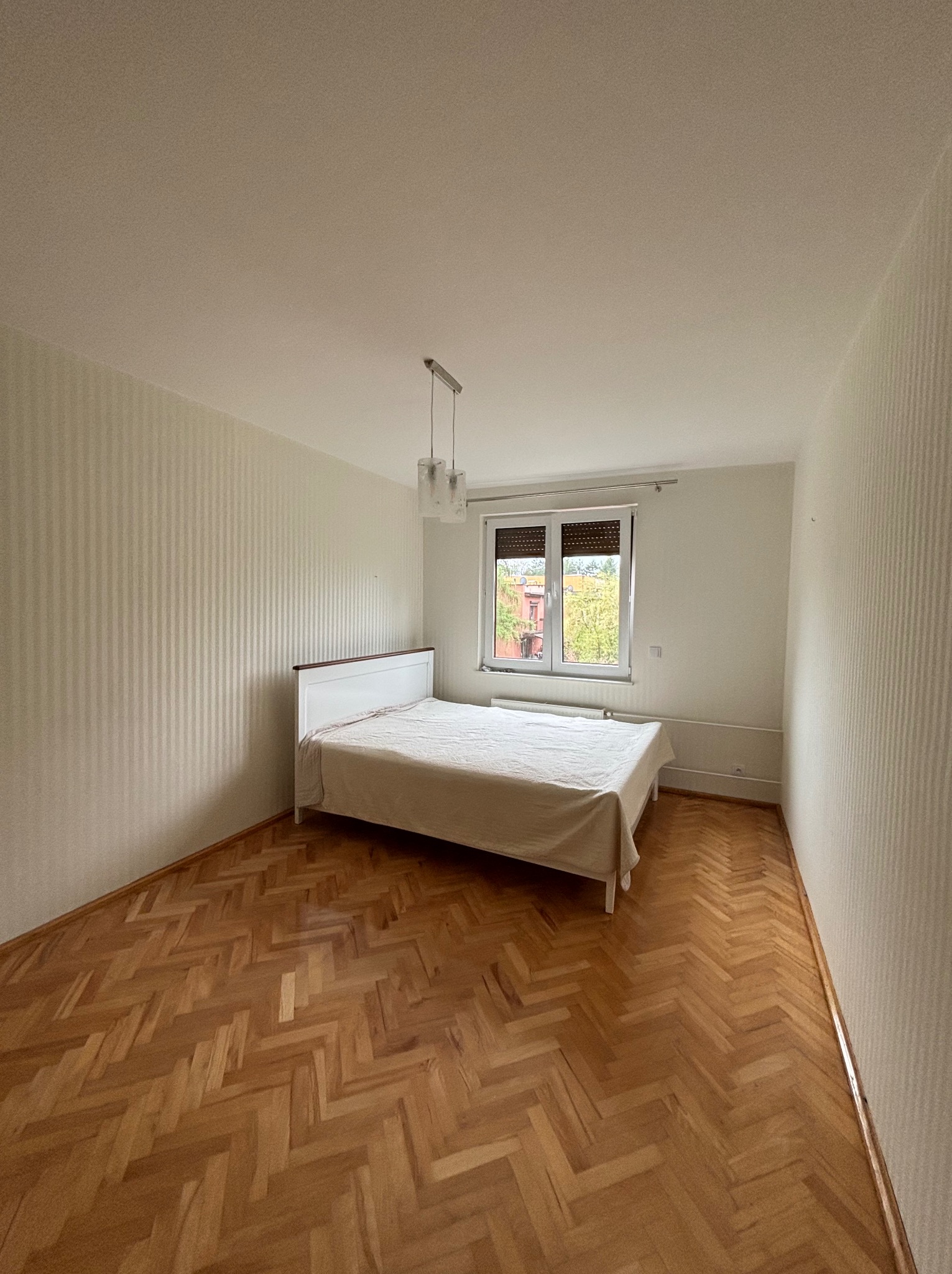 Pokój po remoncie: parkiet w jodełkę, ściany w pionowe pasy, białe łóżko pod oknem z roletami. Widoczne detale wykończenia i oświetlenie.