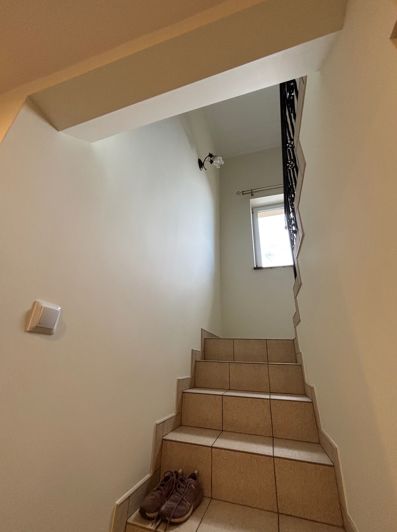 Klatka schodowa z beżowymi płytkami, widok z dołu. Na pierwszym planie para brązowych butów. Ściany w jasnym odcieniu beżu, okno na końcu schodów.