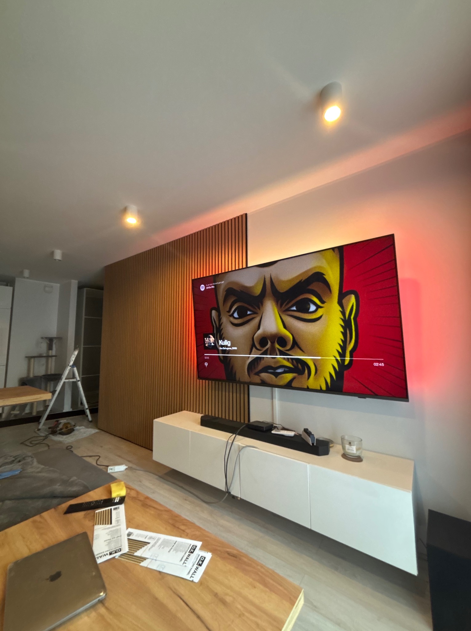 Nowoczesny salon z drewnianą ścianą z lameli, podświetlonym telewizorem i białą szafką RTV. Widoczne narzędzia i materiały sugerują prace wykończeniowe.