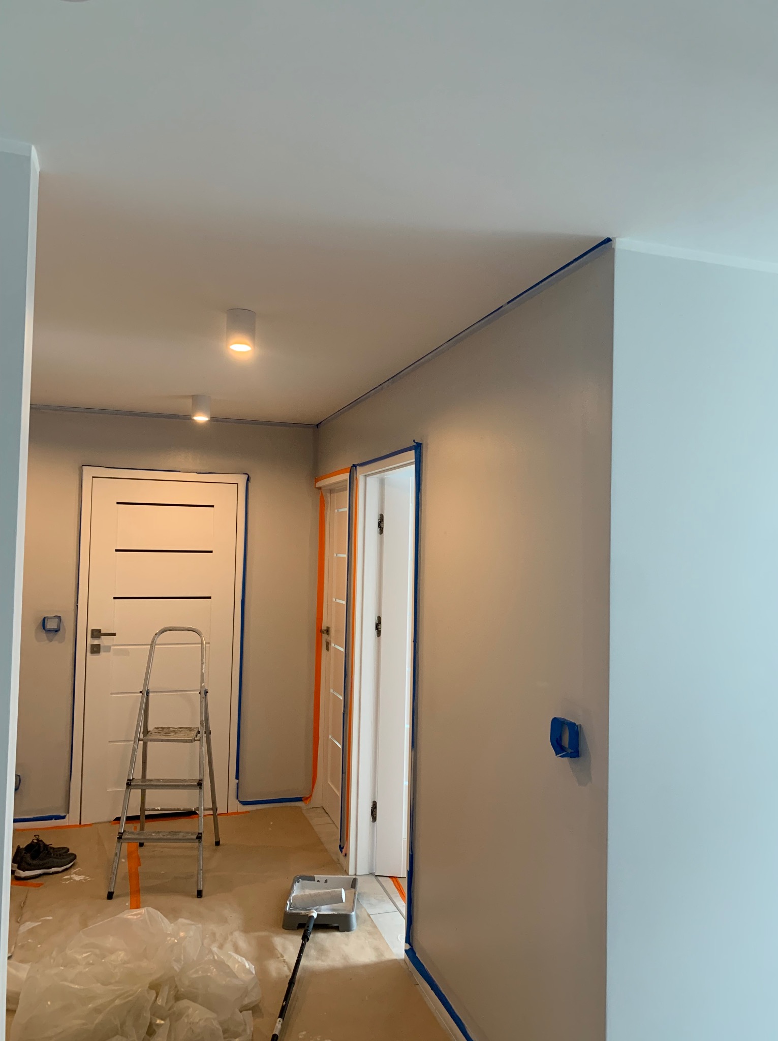Przygotowanie do malowania: zabezpieczone taśmą ściany, drzwi i podłoga. Widoczna drabina, wałek malarski i folia ochronna. Ściany częściowo pomalowane na szaro.