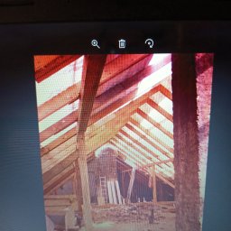 dombud - Konstrukcja dachu z drewnianymi belkami w trakcie budowy, widok z wnętrza poddasza. W tle komin i drabina. Surowe wykończenie, etap prac dekarskich.