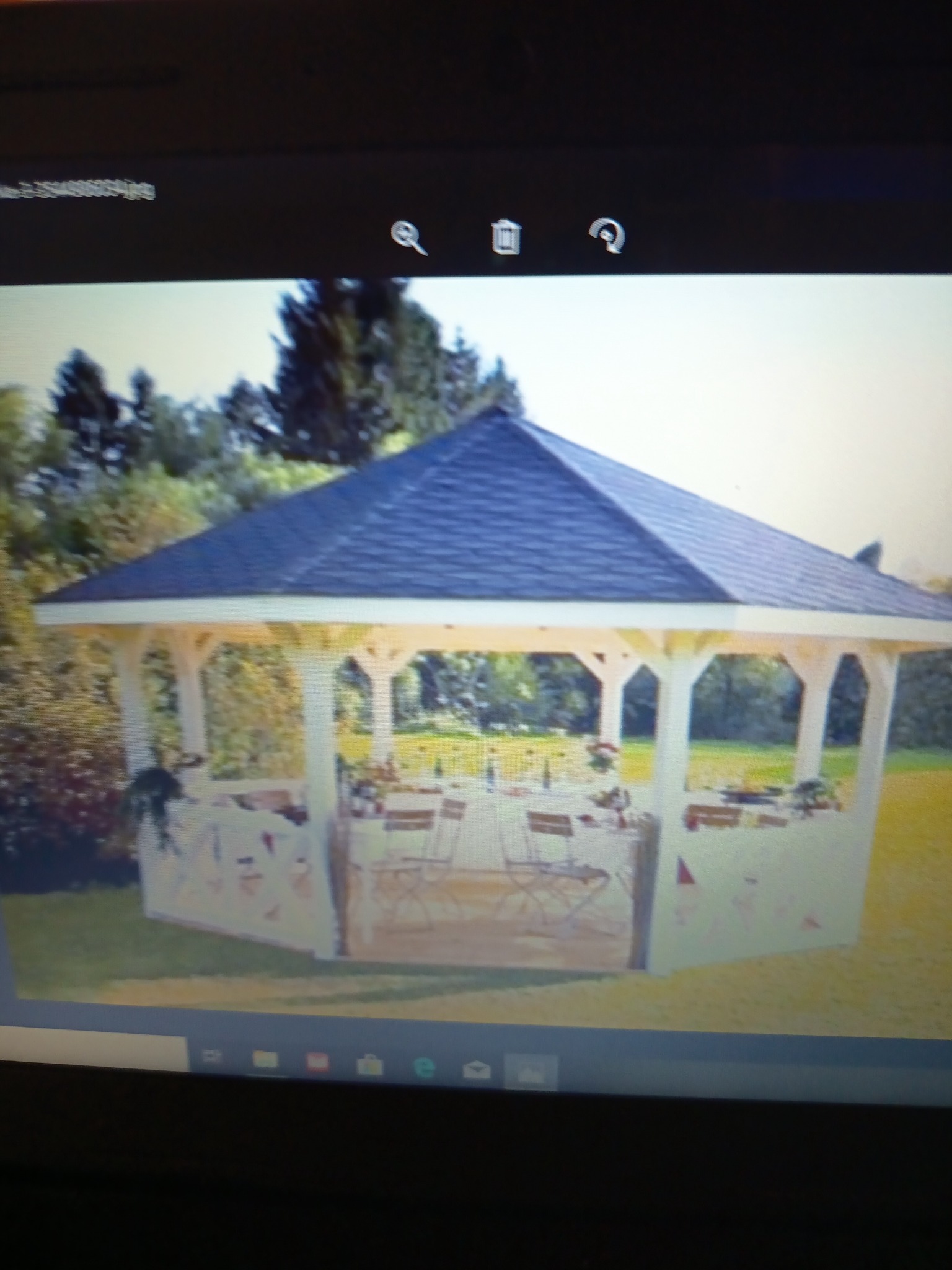 Biała altana z ciemnym dachem, ustawiona na trawniku, z widocznym stołem i krzesłami wewnątrz, otoczona zielenią drzew i krzewów. Widok z zewnątrz.