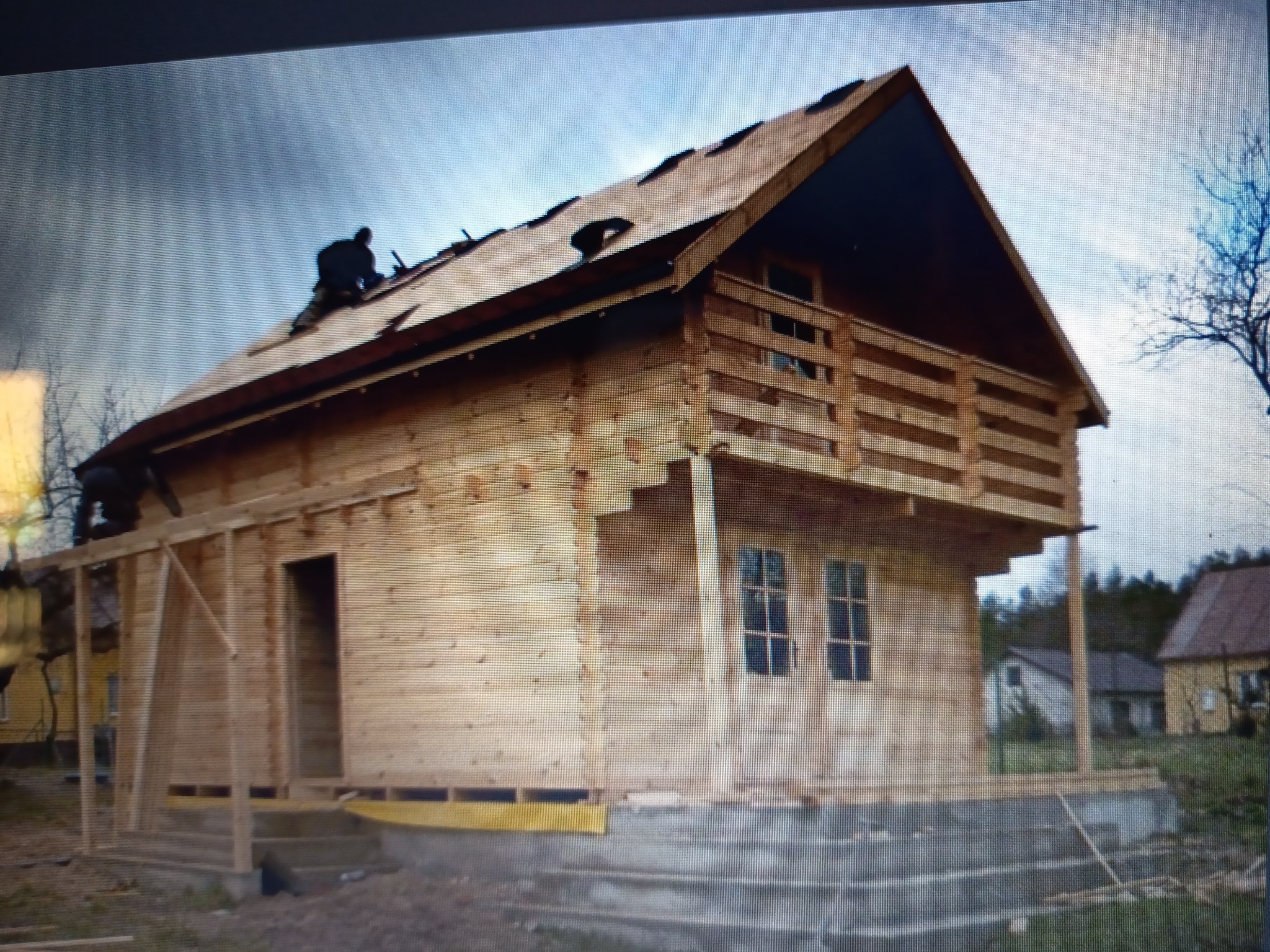 Budowa domu z bali w trakcie realizacji. Widoczni pracownicy na dachu. Surowy stan, brak wykończenia. Ujęcie z dołu, na pierwszym planie fundamenty.