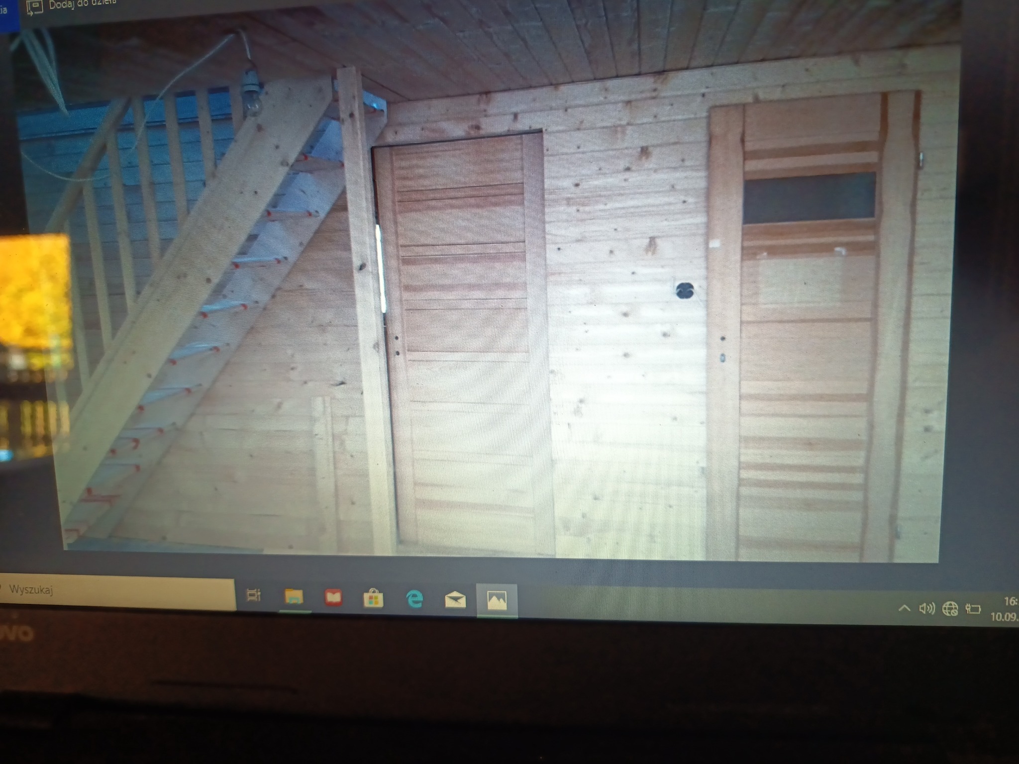 Wnętrze drewnianej konstrukcji z widocznymi schodami drabinkowymi, dwojgiem drzwi, z których jedne mają przeszklenie, wykonane z jasnego drewna.