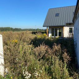 Bartosz Swierczyna - Niedokończony dom z szarego metalu na dachu, otoczony wysoką trawą i chwastami. Widoczne białe ściany z pustaków i drewniane elementy konstrukcyjne.