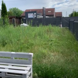 Bartosz Swierczyna - Zarośnięty trawnik z wysoką trawą, widok z perspektywy niskiej, biała paleta meblowa w rogu kadru, szary płot i budynek w tle, pochmurne niebo.