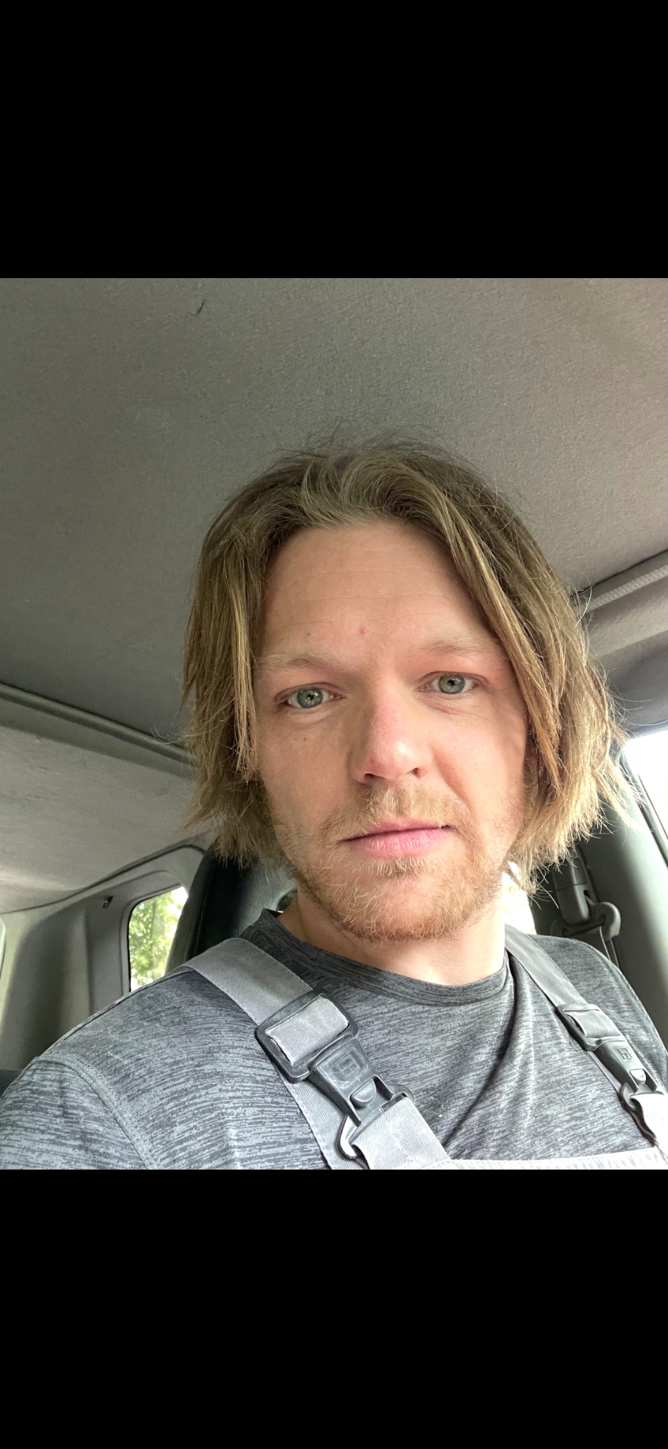 Autoportret mężczyzny z blond włosami i lekkim zarostem, siedzącego w samochodzie z zapiętym pasem bezpieczeństwa. Ujęcie z bliska, naturalne światło.