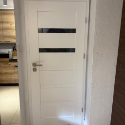 PRO MONTAŻ - Białe drzwi wewnętrzne z poziomymi, czarnymi przeszkleniami, zamontowane w ościeżnicy. W tle widoczna kuchnia z drewnianymi szafkami i fragment podłogi.