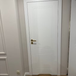 PRO MONTAŻ - Białe, dwuskrzydłowe drzwi z złotą klamką, zamontowane w jasnym wnętrzu. Widoczne listwy przypodłogowe i fragment gniazdka elektrycznego. Na podłodze drobne ślady po montażu.