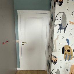 PRO MONTAŻ - Białe drzwi z złotą klamką w pokoju dziecięcym z tapetą w psie wzory. Widoczny fragment szafy i podłogi z paneli. Ujęcie z niskiego kąta.