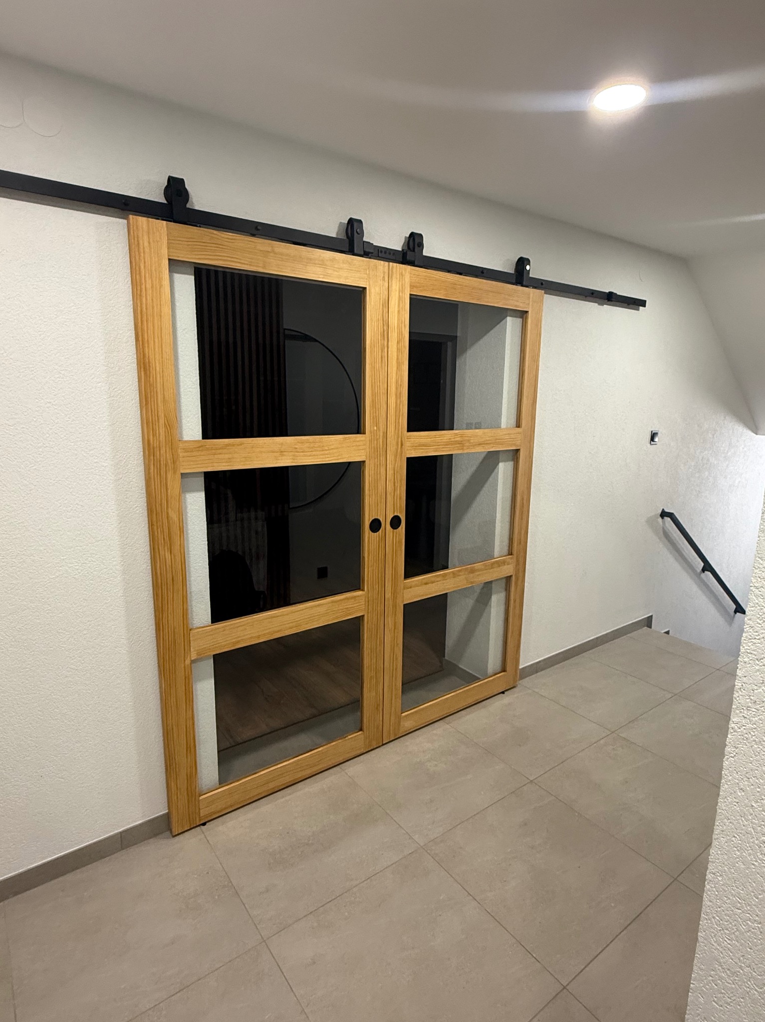 Drewniane, przesuwne drzwi z szybami, zamontowane na czarnej szynie, w jasnym wnętrzu. Widoczne płytki podłogowe i fragment schodów.