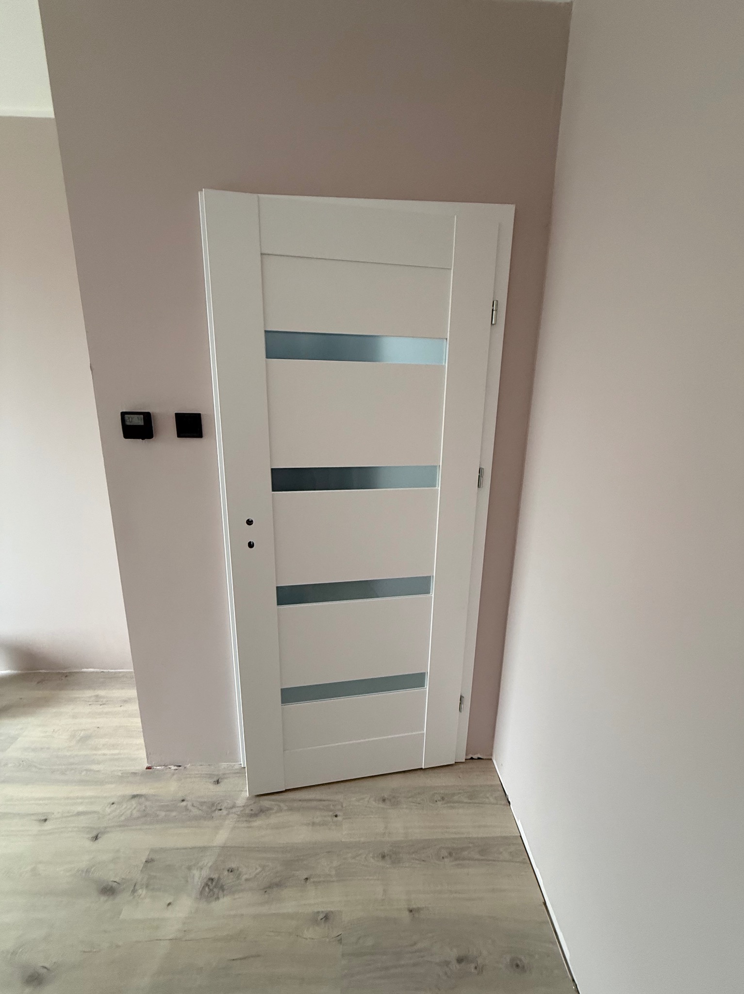 Białe drzwi wewnętrzne z przeszkleniami w trzech rzędach, zamontowane w ścianie pomalowanej na jasny róż, z widoczną podłogą z paneli imitujących drewno.