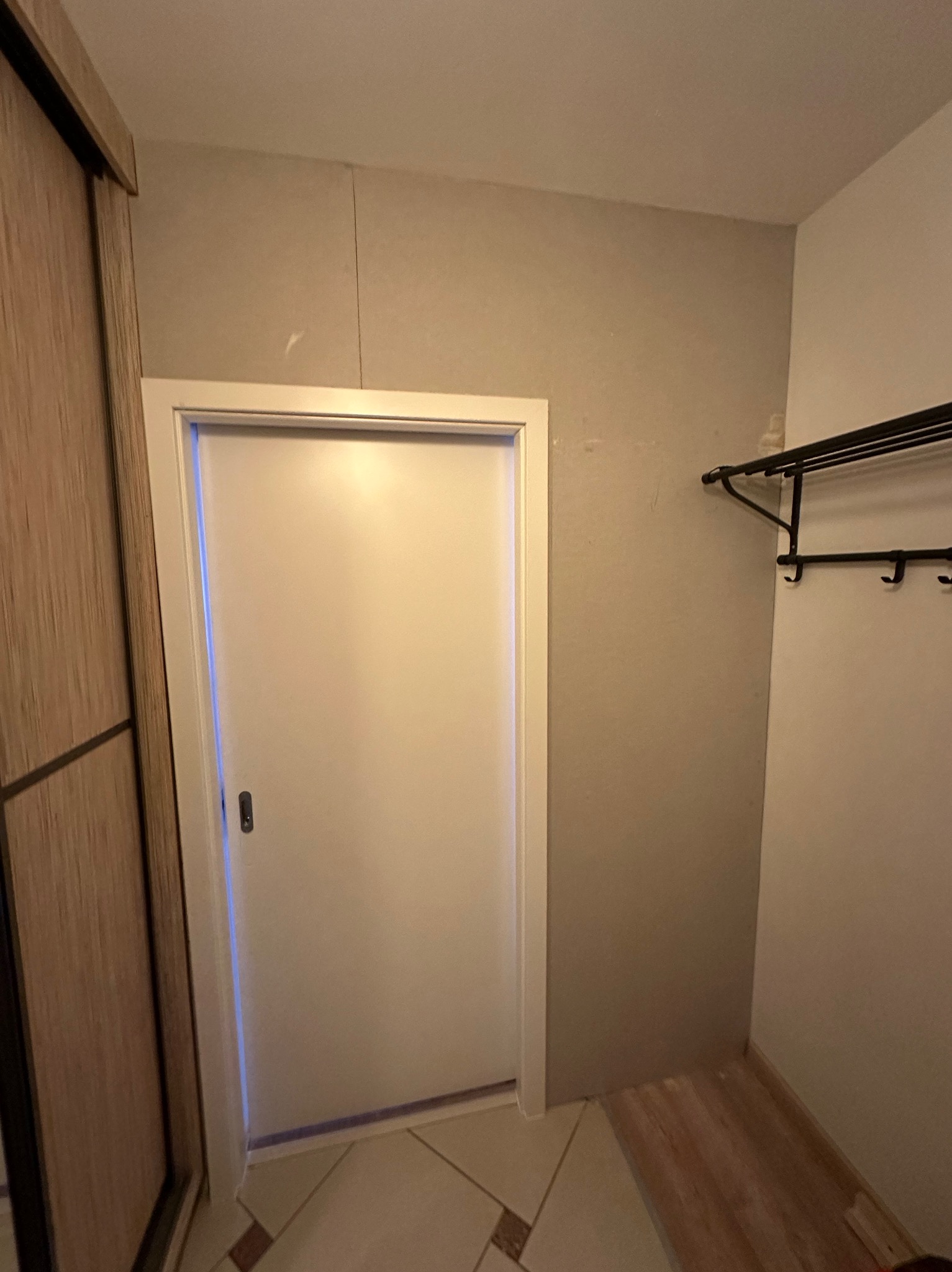 Białe drzwi wewnętrzne z ościeżnicą, widoczne z bliska w korytarzu, obok szafy przesuwnej i wieszaka na ubrania. Minimalistyczny design, neutralna kolorystyka.