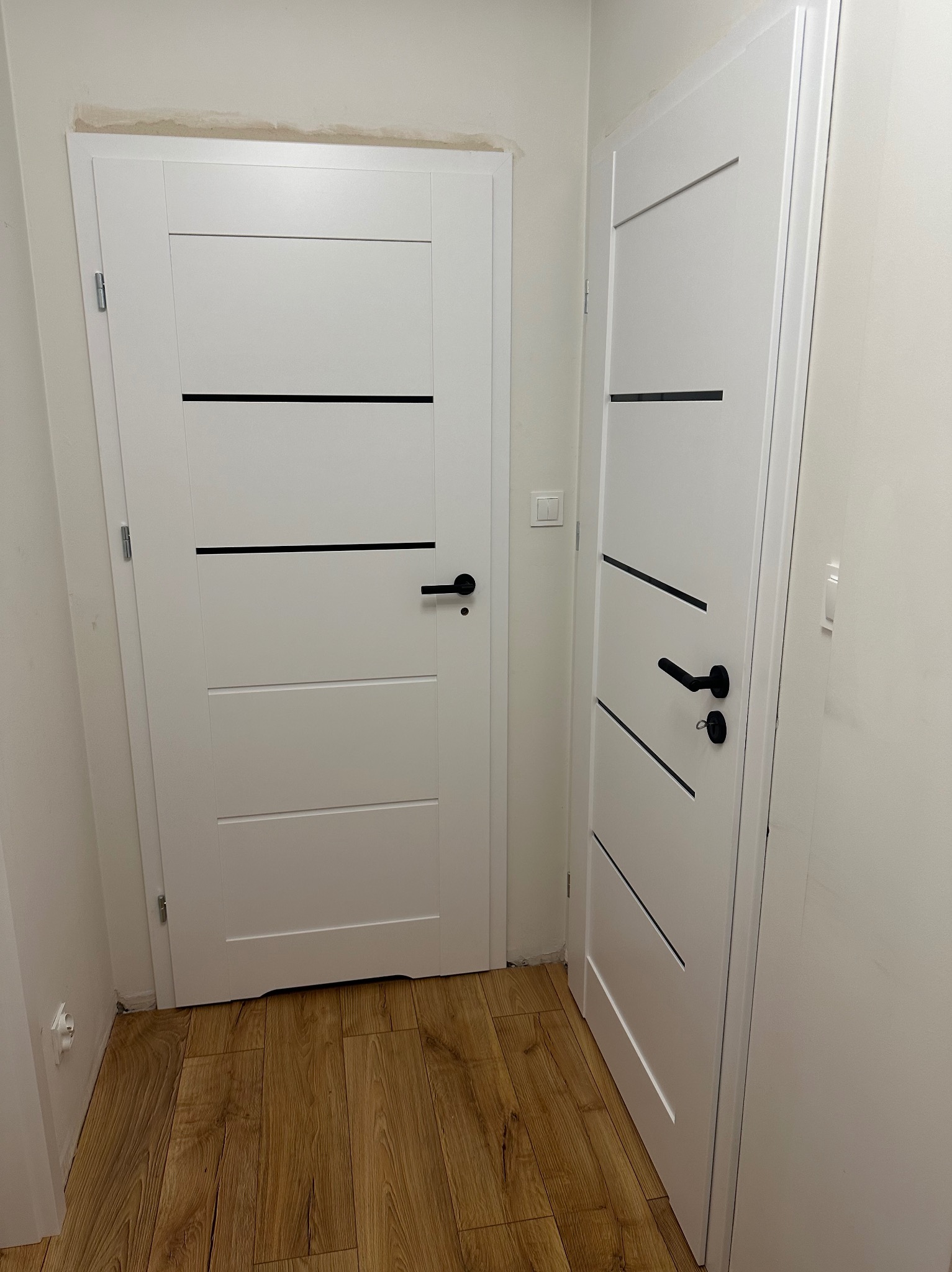 Dwie białe, nowoczesne drzwi wewnętrzne z czarnymi, poziomymi akcentami i klamkami na tle parkietu. Widoczne niedoskonałości wykończenia przy futrynie.