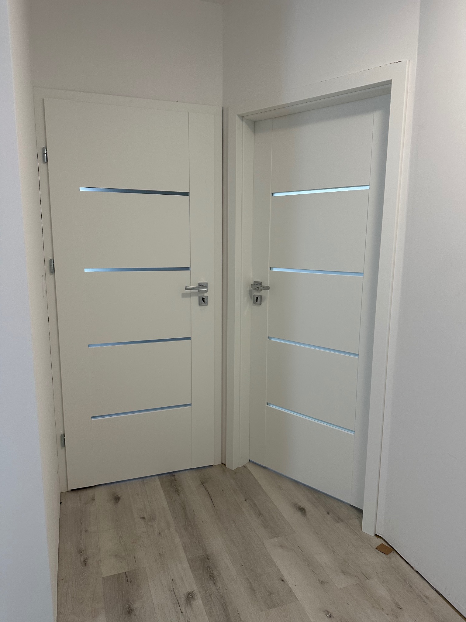 Dwa białe, nowoczesne drzwi wewnętrzne z poziomymi przeszkleniami, zamontowane w jasnym pomieszczeniu z panelami podłogowymi. Widok z perspektywy podłogi.