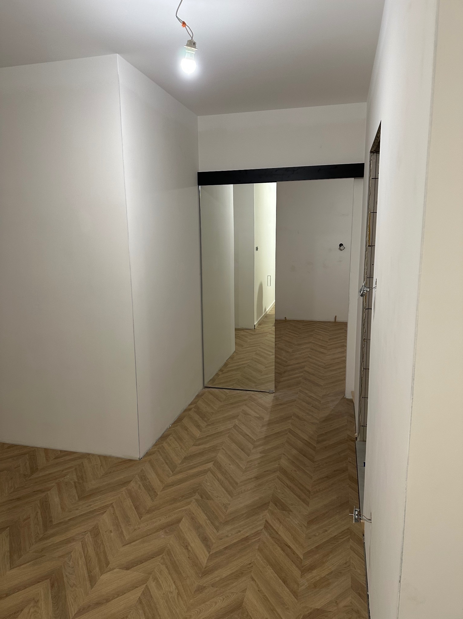 Nowoczesne wnętrze z ułożoną podłogą w jodełkę, widok na przesuwne drzwi z czarną ramą i surową ścianę w trakcie wykończenia. Minimalistyczny design.