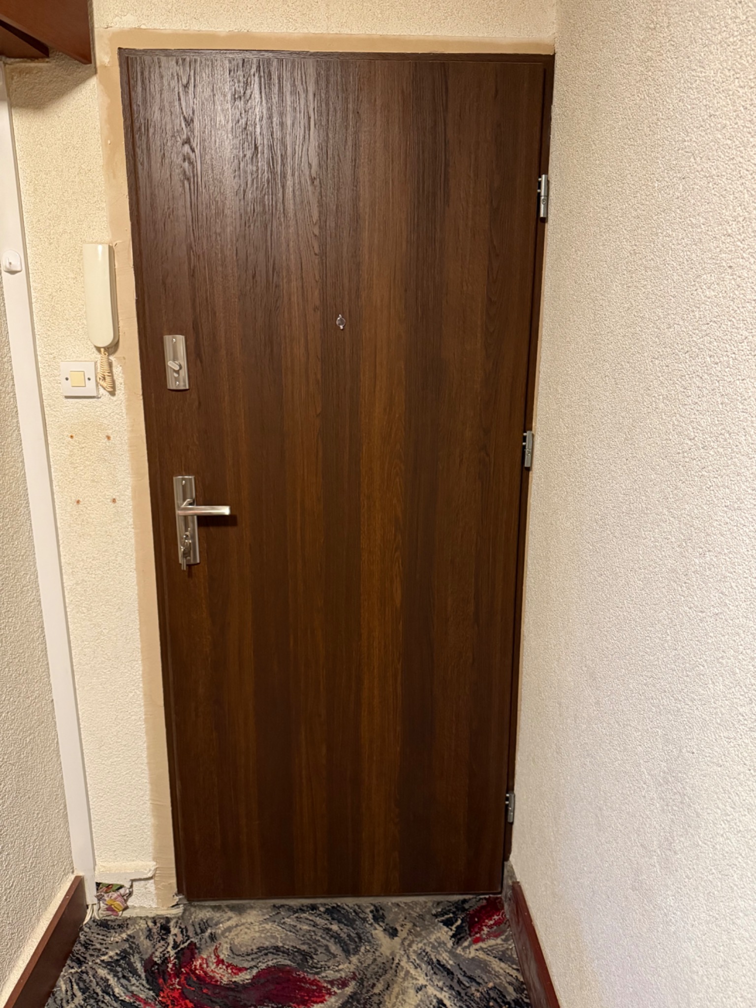 Ciemnobrązowe drzwi wejściowe z wizjerem i srebrną klamką, zamontowane w jasnym przedpokoju z beżowymi ścianami i wzorzystą wykładziną podłogową. Widok z bliska.