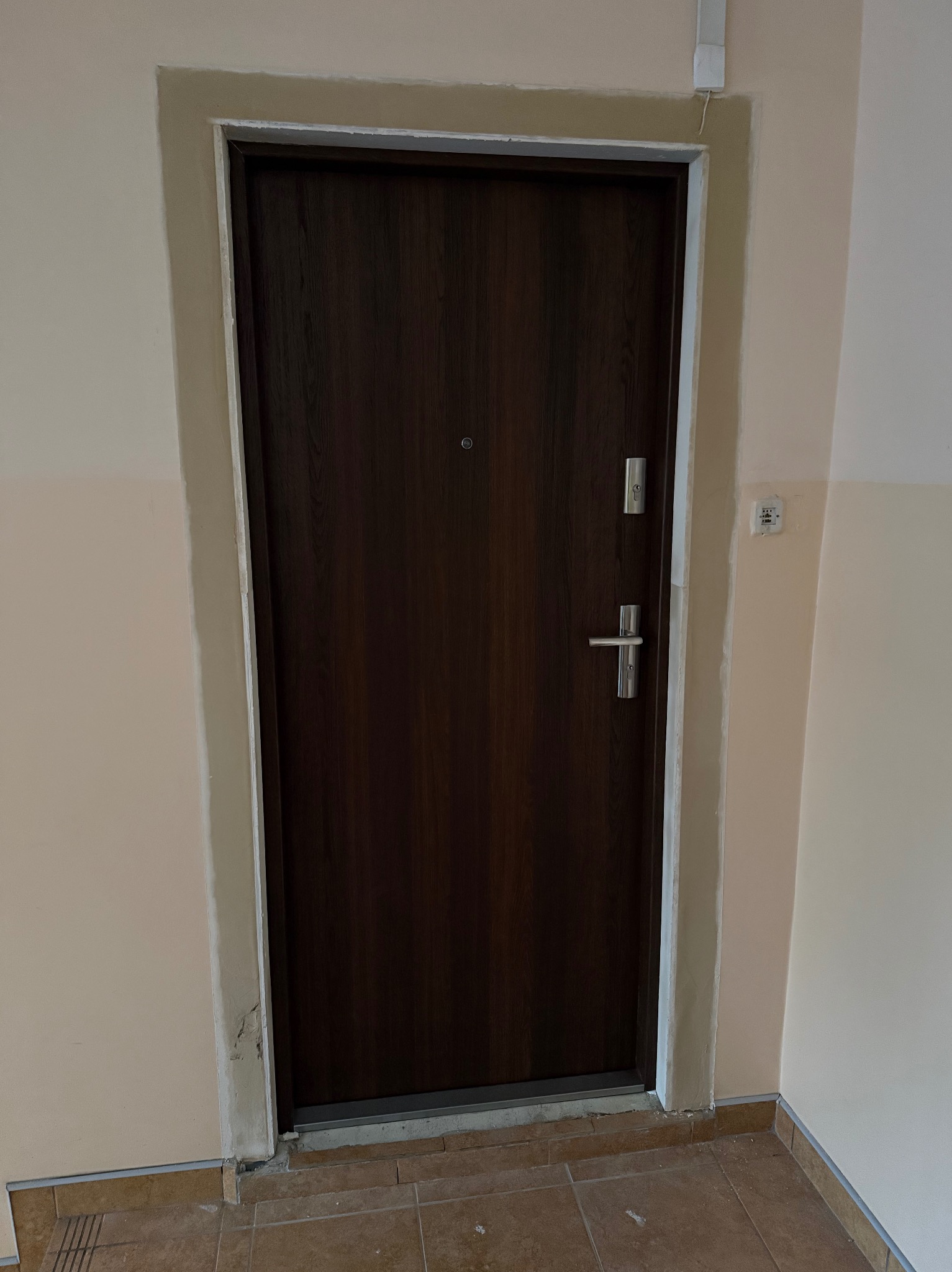 Ciemne drzwi wejściowe z wizjerem i srebrną klamką, osadzone w beżowej futrynie, na tle ściany w odcieniach beżu i brązu. Widoczny fragment podłogi z płytek.