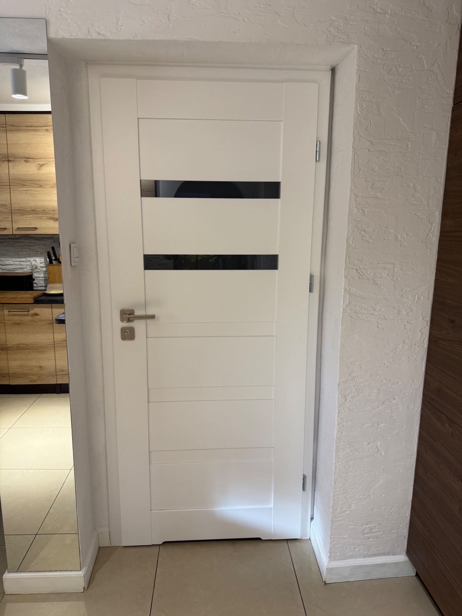 Białe drzwi wewnętrzne z poziomymi, czarnymi przeszkleniami, zamontowane w ościeżnicy. W tle widoczna kuchnia z drewnianymi szafkami i fragment podłogi.