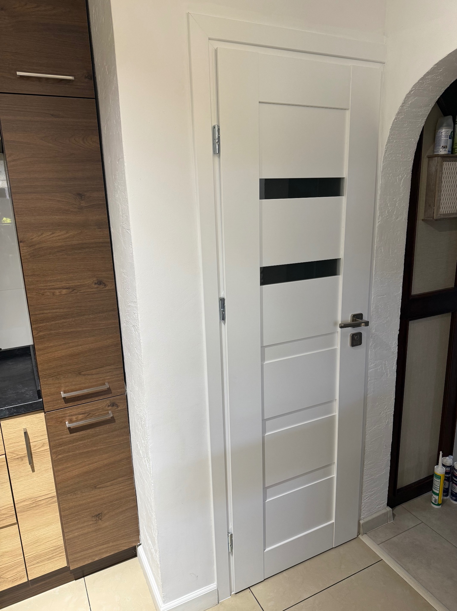 Białe, nowoczesne drzwi wewnętrzne z poziomymi, czarnymi paskami, zamontowane w białej ścianie, widoczne obok drewnianej szafki kuchennej.