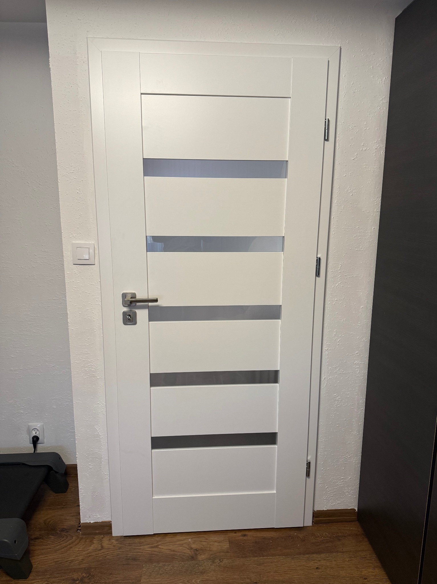 Białe drzwi wewnętrzne z czterema poziomymi przeszkleniami, zamontowane w ścianie o nierównej fakturze. Widoczny fragment podłogi i szafy.