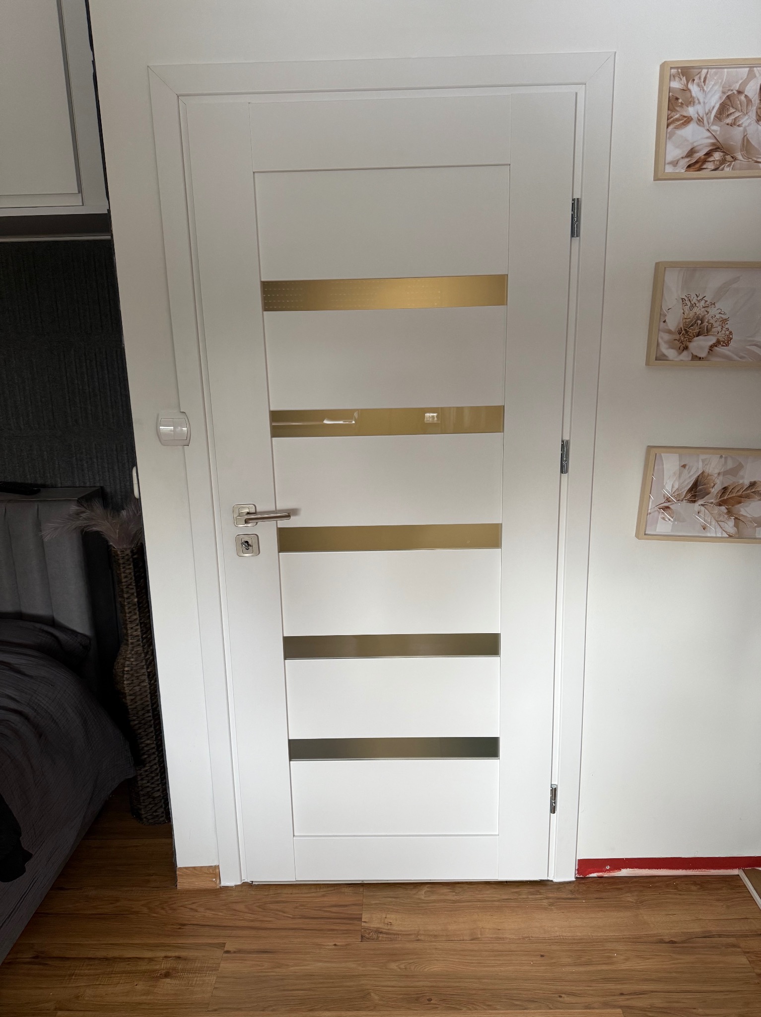 Białe drzwi wewnętrzne z poziomymi, złotymi paskami, zamontowane w pokoju z panelami podłogowymi i dekoracjami ściennymi w beżowych ramkach.