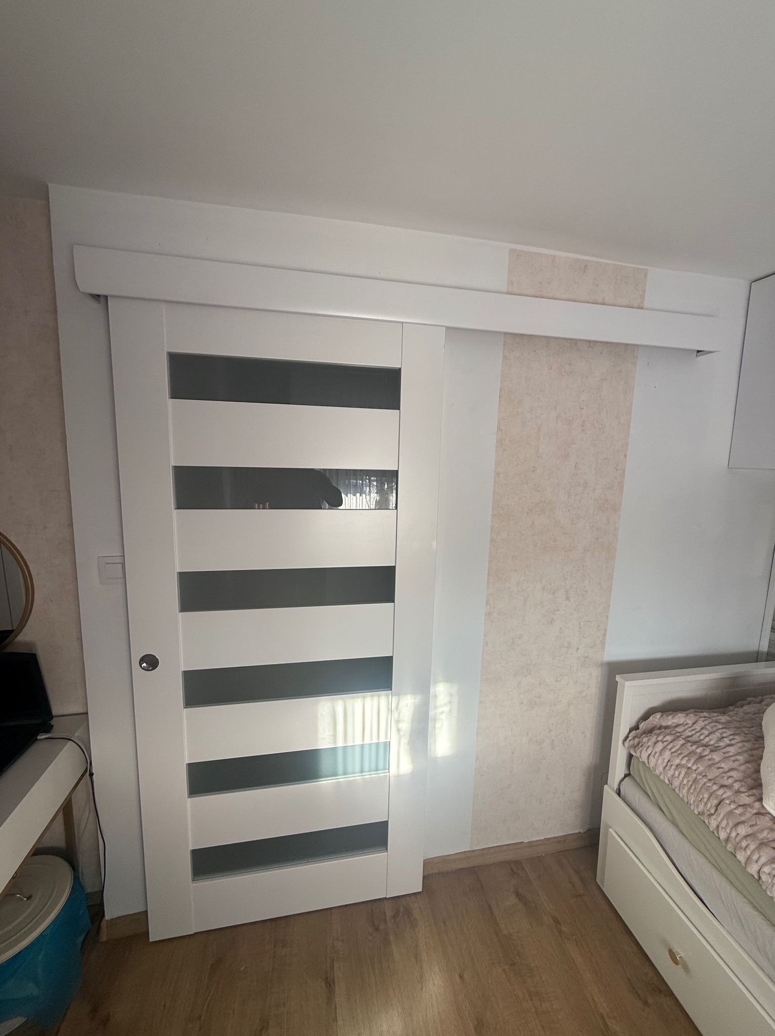 Białe, przesuwne drzwi z poziomymi, szklanymi pasami, zamontowane w pokoju z beżową tapetą. Widoczny fragment łóżka i szafki nocnej.