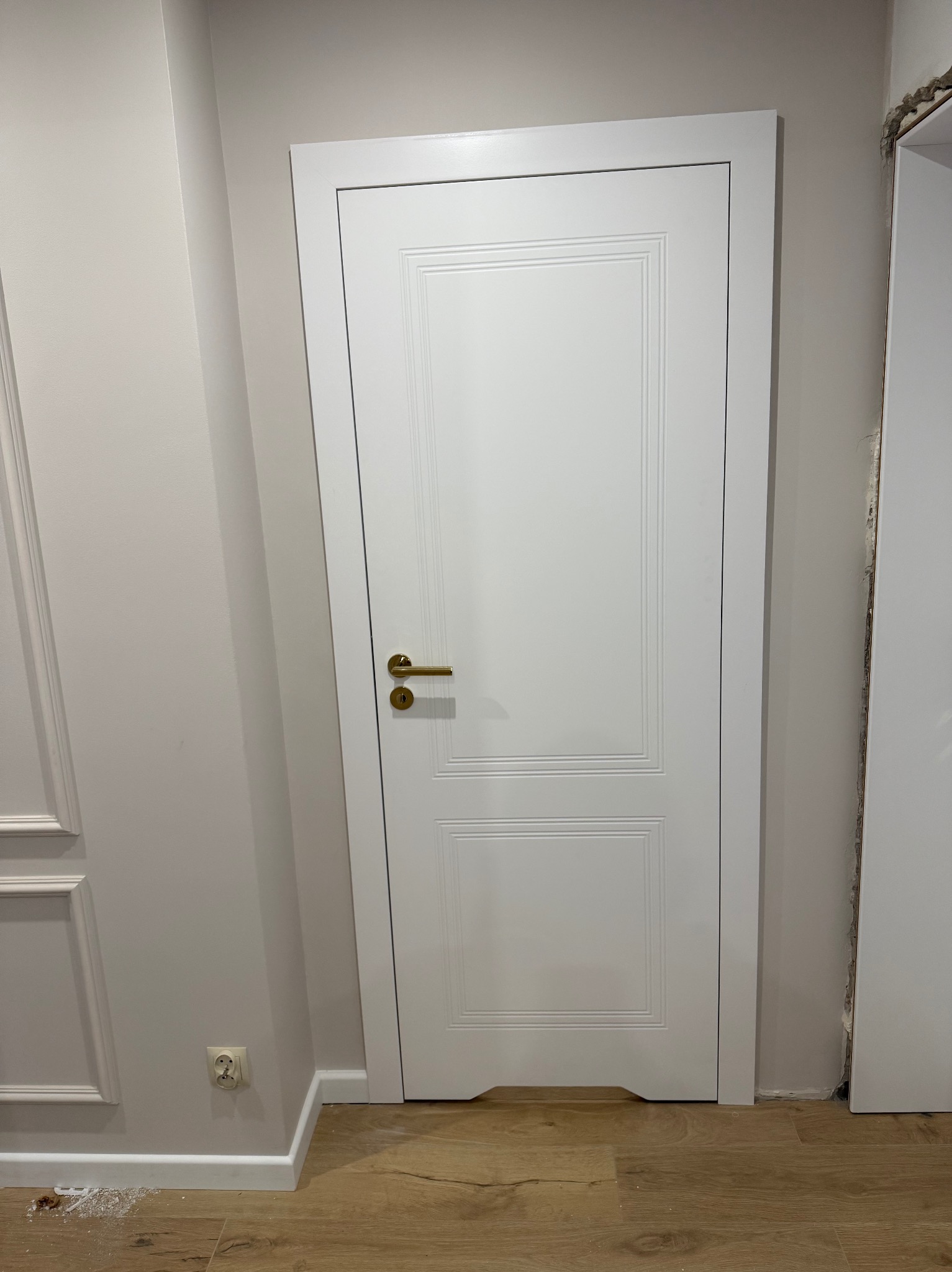 Białe, dwuskrzydłowe drzwi z złotą klamką, zamontowane w jasnym wnętrzu. Widoczne listwy przypodłogowe i fragment gniazdka elektrycznego. Na podłodze drobne ślady po montażu.