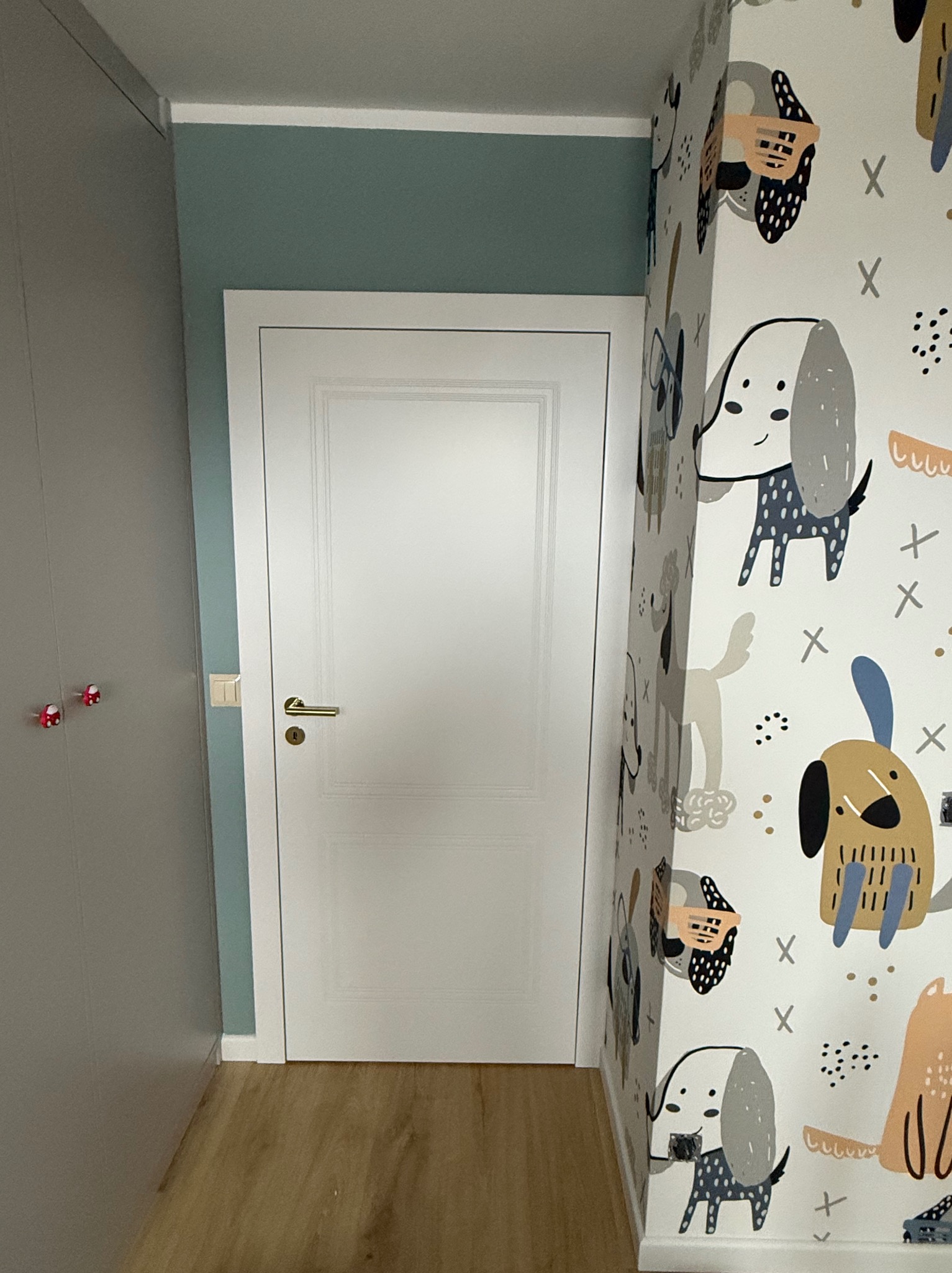 Białe drzwi z złotą klamką w pokoju dziecięcym z tapetą w psie wzory. Widoczny fragment szafy i podłogi z paneli. Ujęcie z niskiego kąta.