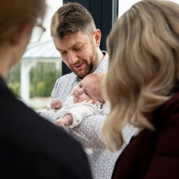 Tomasz Gościewicz Fotografia - Tata trzymający niemowlę na rękach, z uśmiechem patrzy na dziecko. W tle widoczne dwie osoby, prawdopodobnie rodzina, obserwujące tę wzruszającą chwilę.