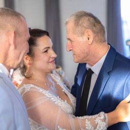 Tomasz Gościewicz Fotografia - Uśmiechnięta panna młoda w koronkowej sukni spogląda na pana młodego w granatowym garniturze. W tle fotografka z aparatem.