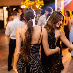 Tomasz Gościewicz Fotografia - Dwie uśmiechnięte kobiety w błyszczących sukienkach na parkiecie podczas eventu. W tle rozmyte postacie i dekoracje świetlne.