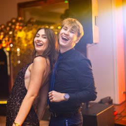 Tomasz Gościewicz Fotografia - Uśmiechnięta para tańczy na parkiecie podczas eventu. Kobieta w cekinowej sukience, mężczyzna w koszuli i krawacie. W tle dekoracje świetlne i nagłośnienie.