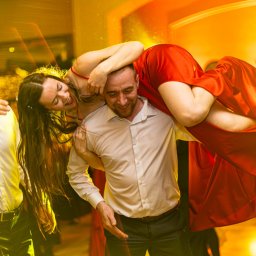 Tomasz Gościewicz Fotografia - Radosna zabawa weselna w Głogowie: mężczyzna trzyma na ramionach uśmiechniętą kobietę w czerwonej sukni, otoczonych przez rozbawionych gości w ciepłym oświetleniu.