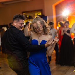 Tomasz Gościewicz Fotografia - Para tańczy na parkiecie podczas eventu. Kobieta w niebieskiej sukience, mężczyzna w czarnej koszuli. W tle rozświetlona scena i goście.